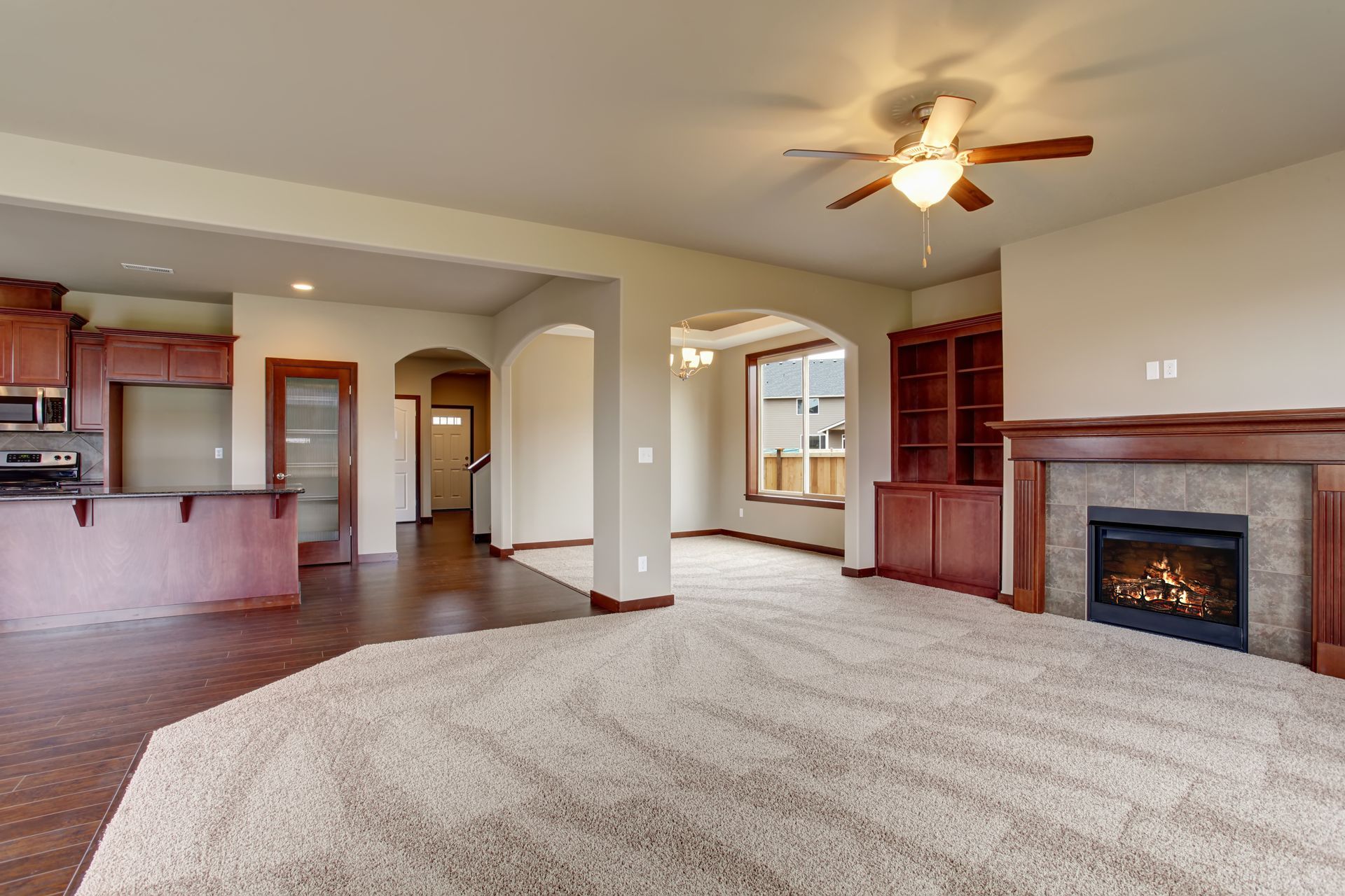 Empty living room with beige carpet, fireplace, ceiling fan, and open kitchen area Empty living room with beige carpet, fireplace, ceiling fan, and open kitchen area