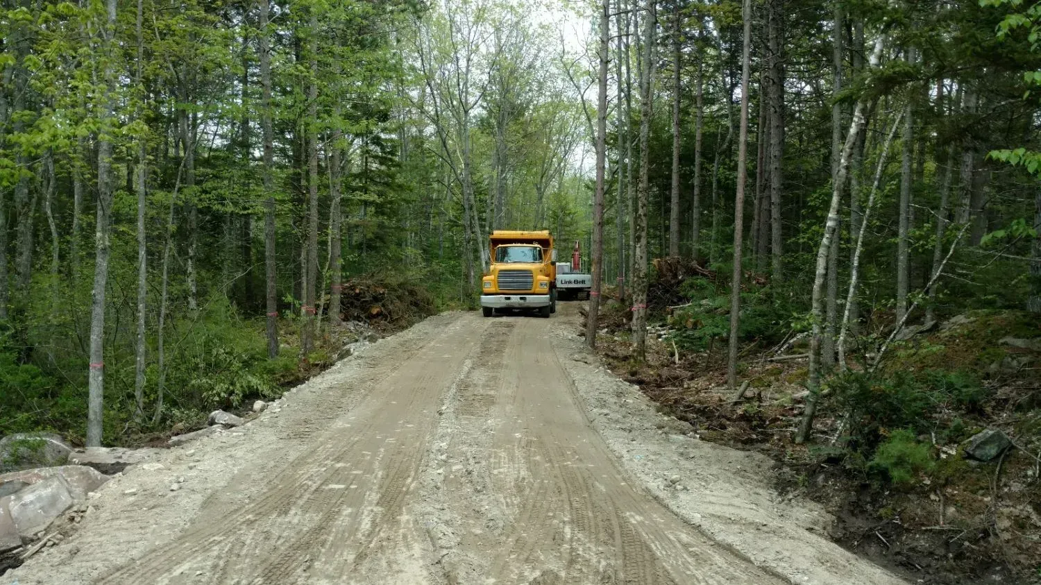 A yellow truck is driving down a dirt road in the woods.