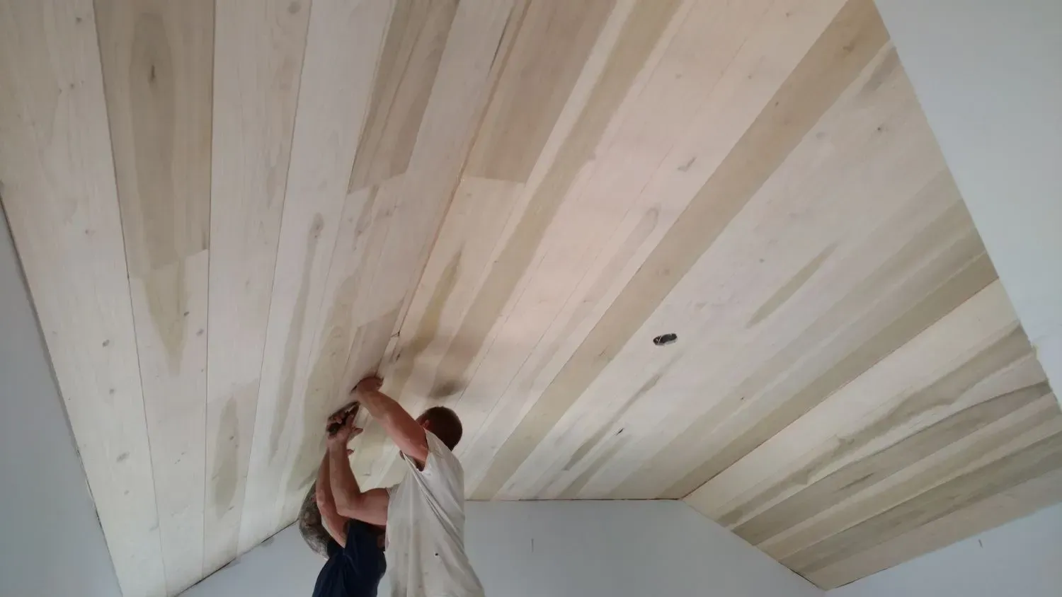 Two men are working on a wooden ceiling in a room.