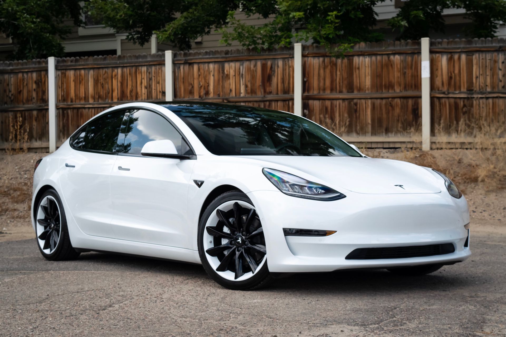 A white tesla model 3 is parked in front of a wooden fence.