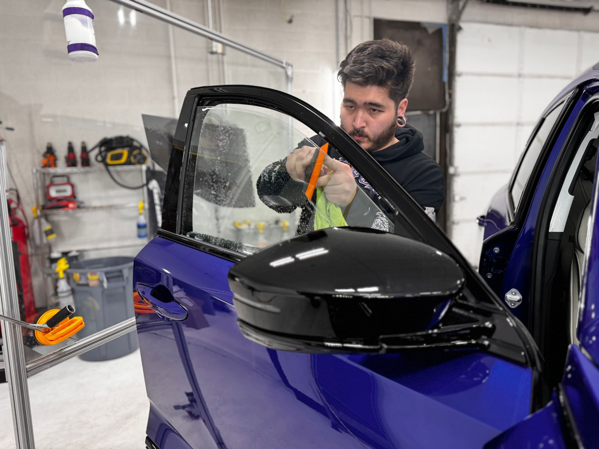 A man is applying window tinting to a blue car in a garage.