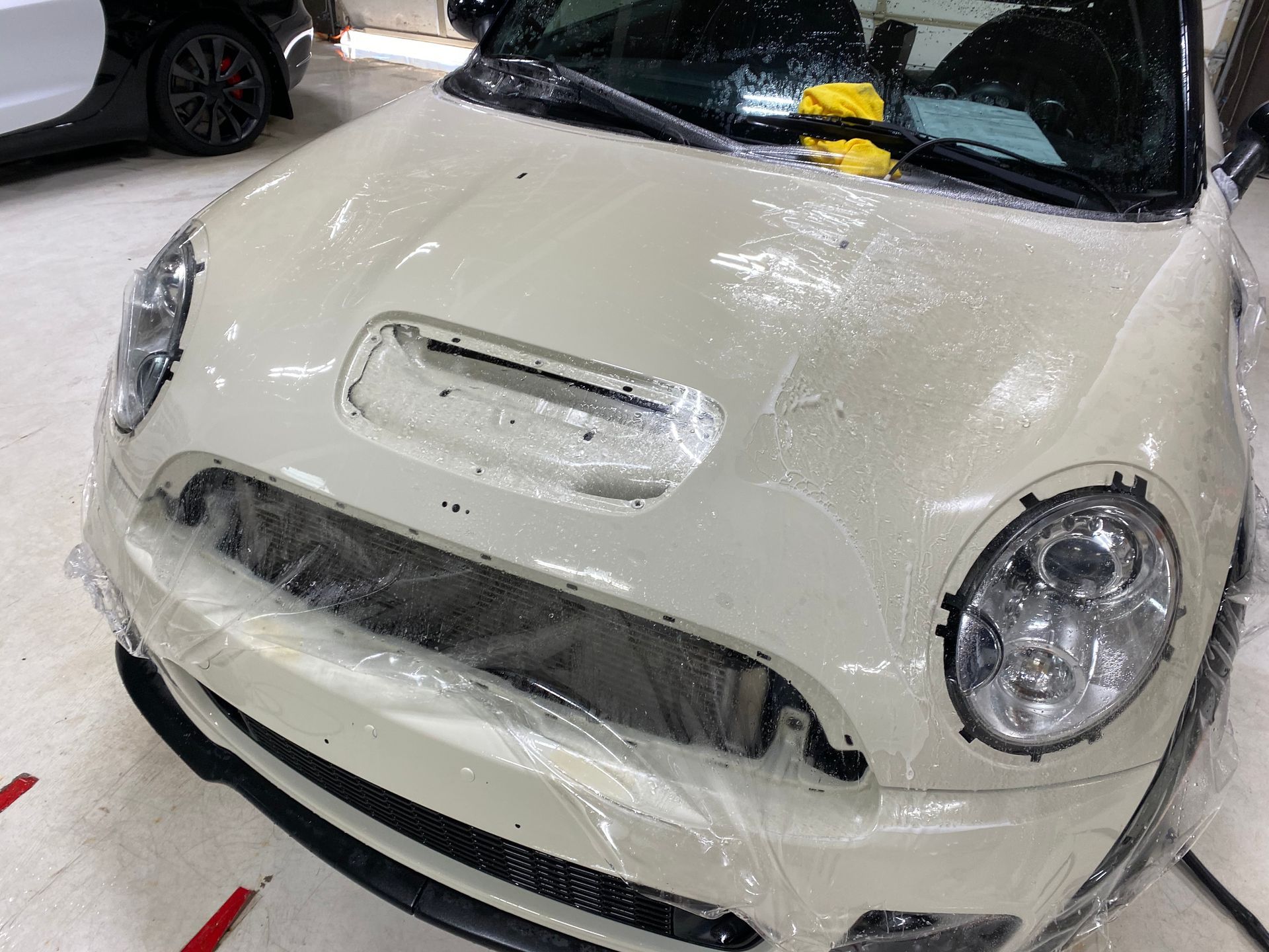 A white mini cooper is covered in plastic wrap in a garage.