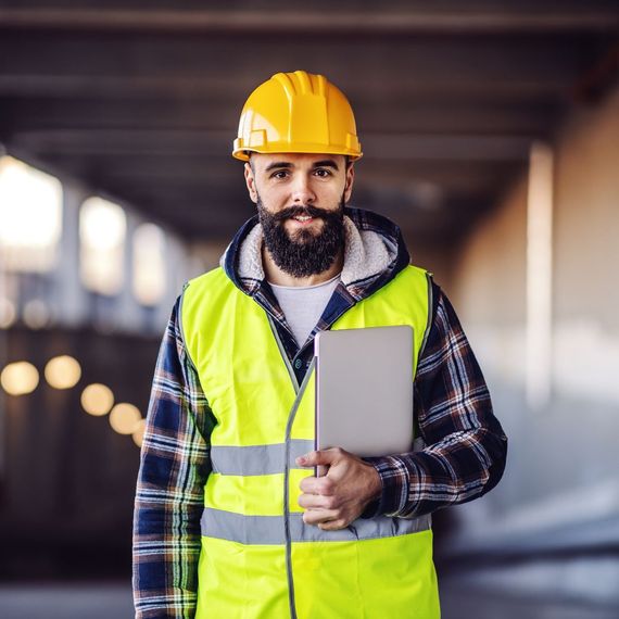 A man wearing a hard hat and a yellow vest is holding a laptop.