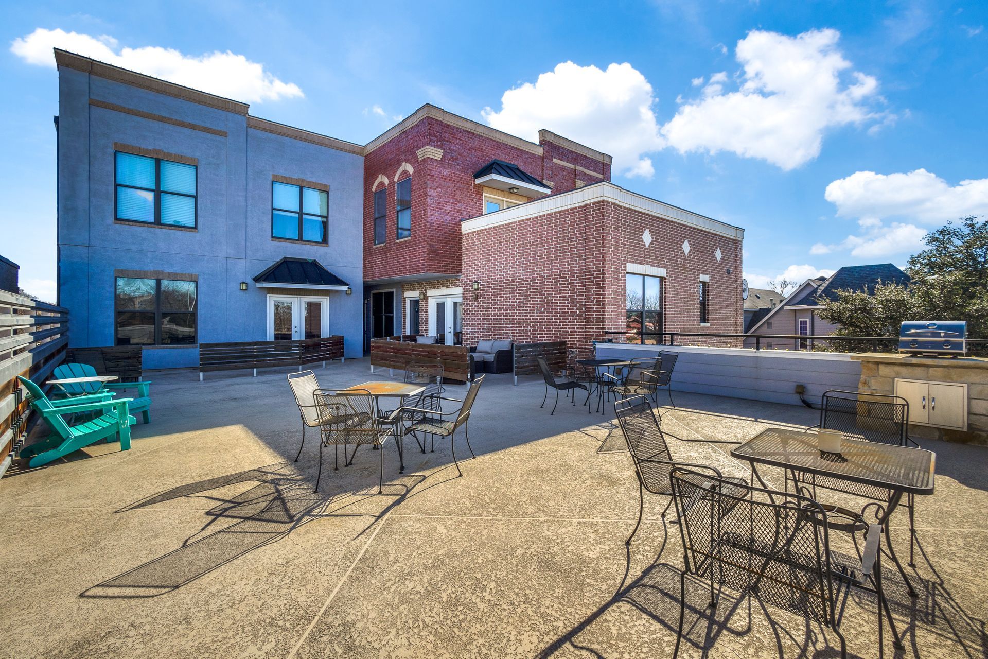 Rooftop patio with tables, chairs, and building with blue, red, and brown brick facades under a blue sky.