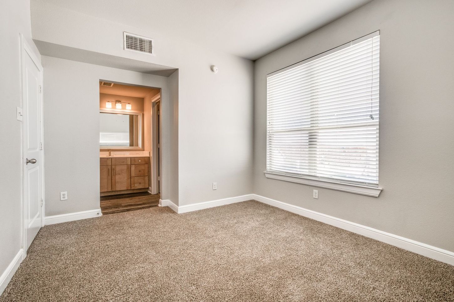 Empty bedroom with beige carpet, window, and doorway to a bathroom.