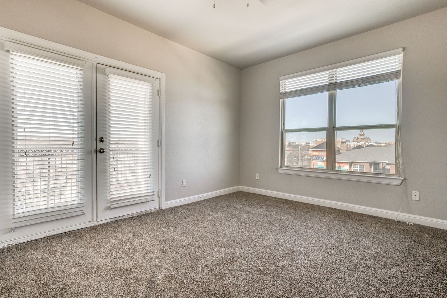 Empty bedroom with closed French doors, window with blinds, beige carpet, and gray walls.