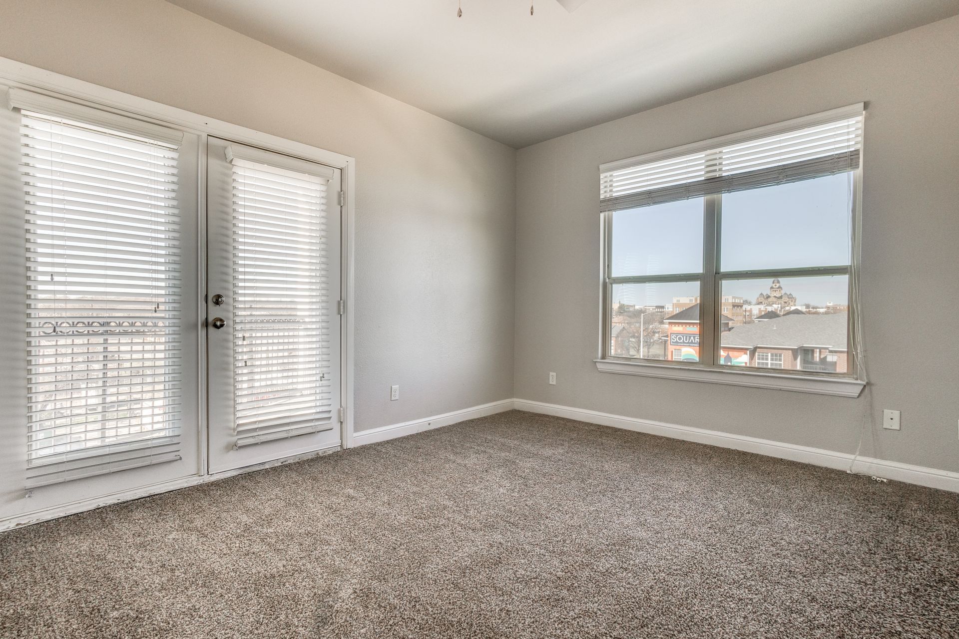 Empty bedroom with closed French doors, window with blinds, beige carpet, and gray walls.