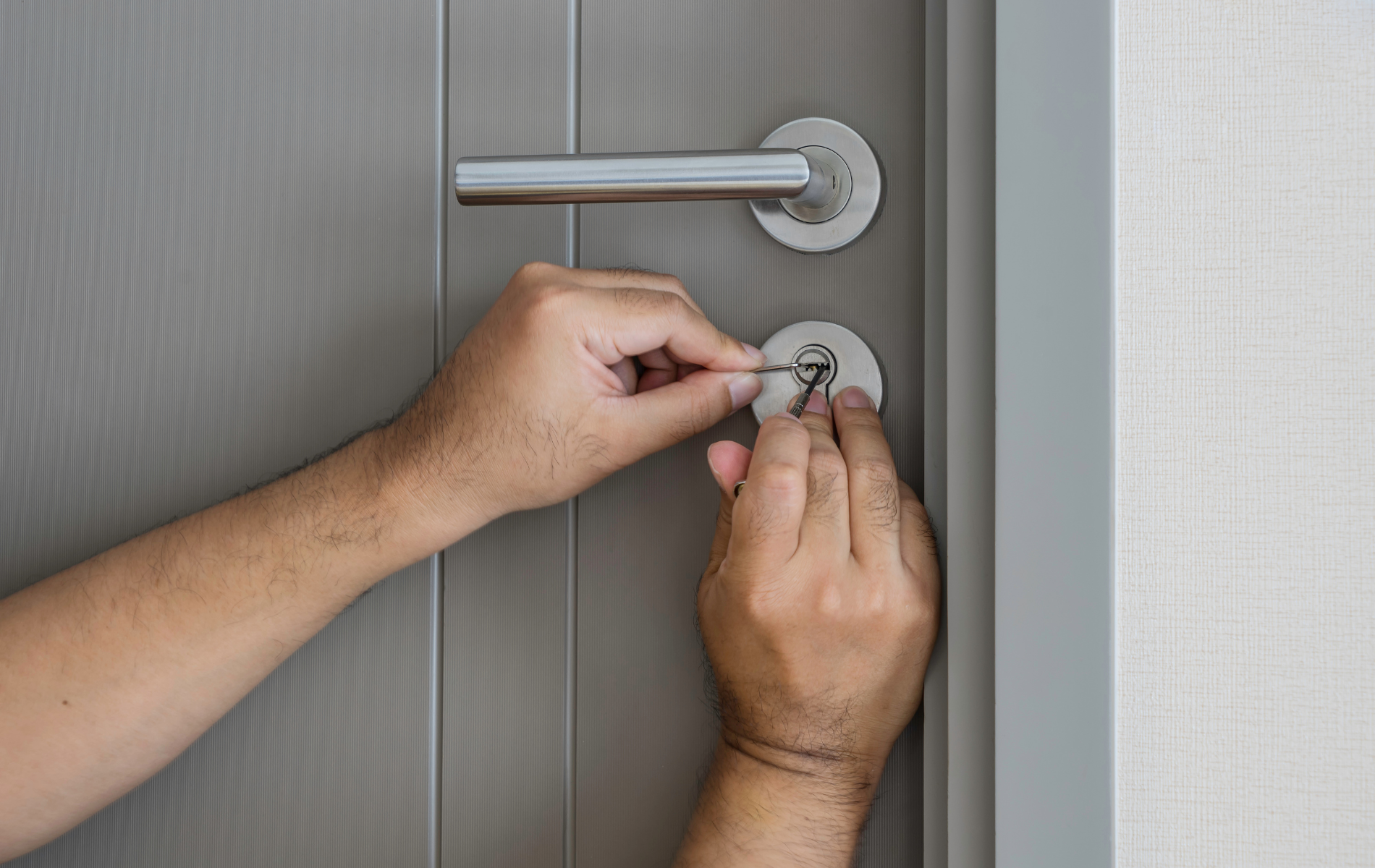 Hands picking a door lock with a pick. Gray door, silver handle and lock, white wall.