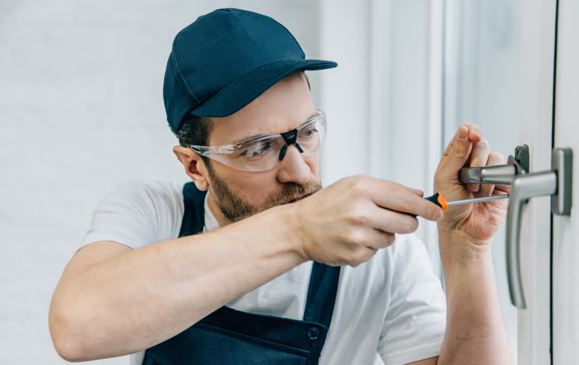 Person in a blue cap and overalls, wearing safety glasses, uses a screwdriver on a window handle.