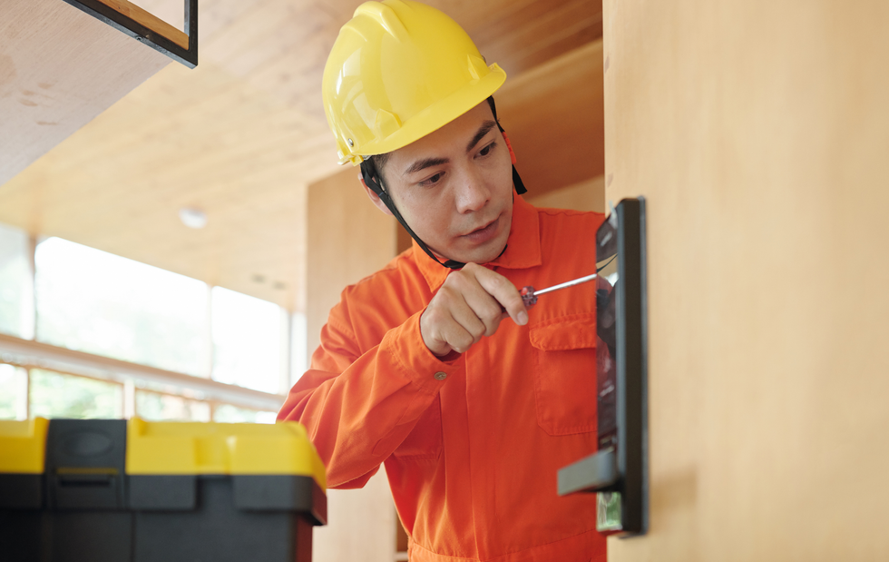 Construction worker in orange overalls and yellow hard hat using a screwdriver on a door lock.
