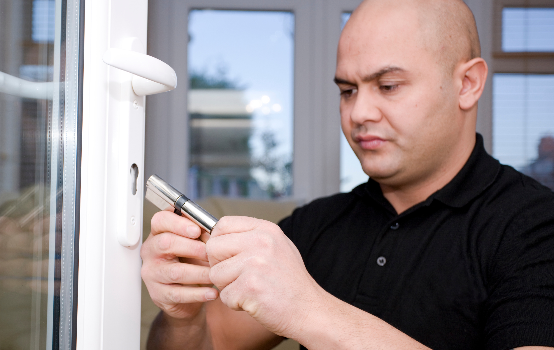 Bald man with furrowed brow using a tool to unlock a white door with white window frame.