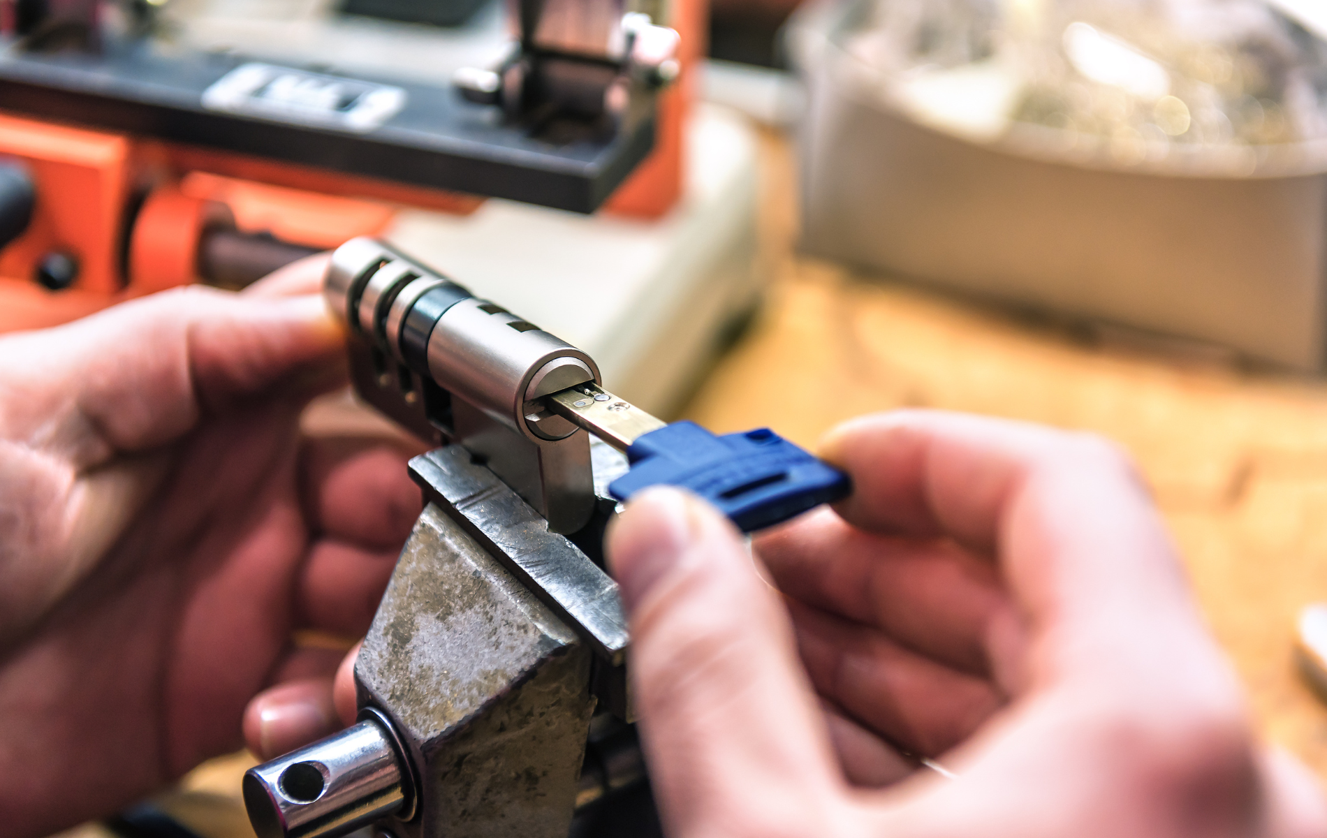 Hands holding a key and lock cylinder in a vise, locksmithing. Key is blue, setting is workshop.