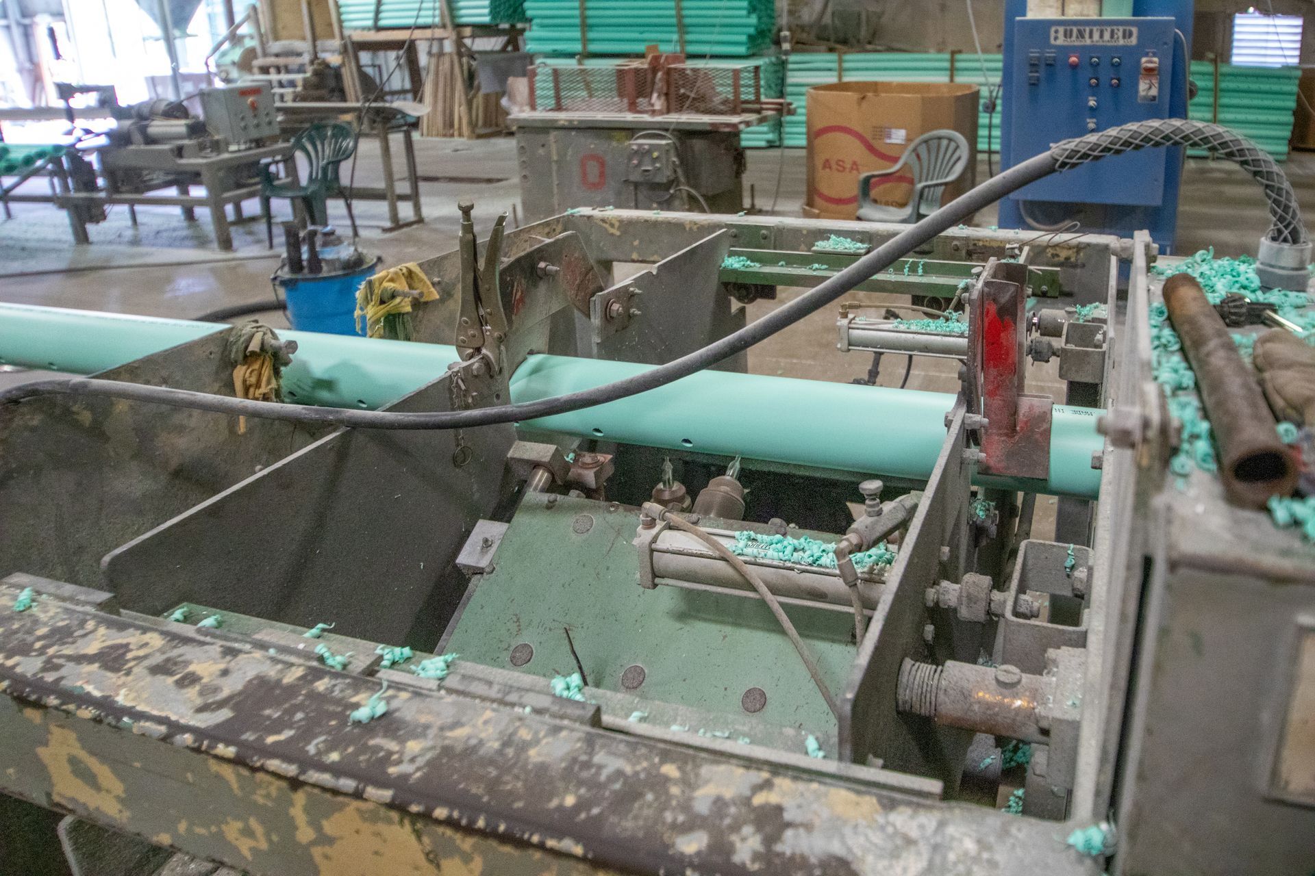 Industrial machine cutting green pipe in a workshop; debris scattered.