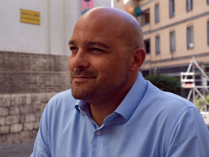 Un uomo calvo con una camicia blu sorride davanti a un edificio