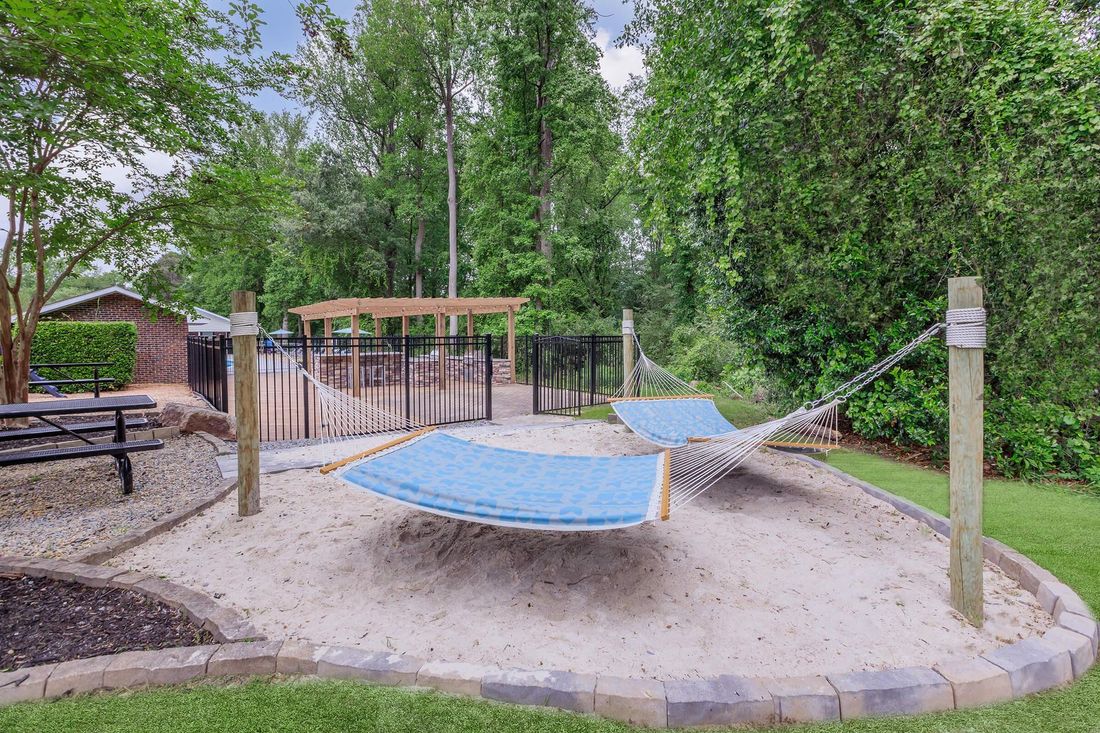 Hammocks in sandy area, bordered by stones, with trees and a fence in the background.