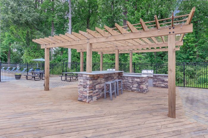 Wooden pergola over a stone bar and grill area on a sandy patio; picnic tables and fence in background.