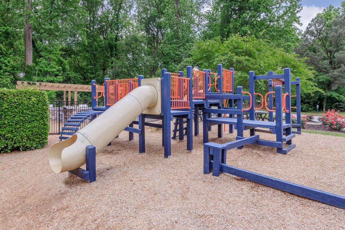 Playground with a beige slide, blue structure, and orange accents, set on wood chips, surrounded by greenery.
