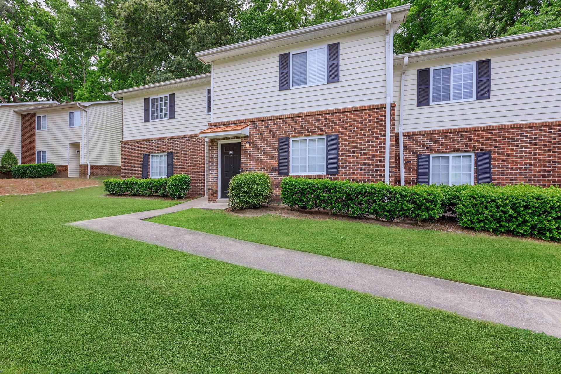Apartment complex exterior, two-story brick and siding buildings, green lawn and bushes, paved walkway.