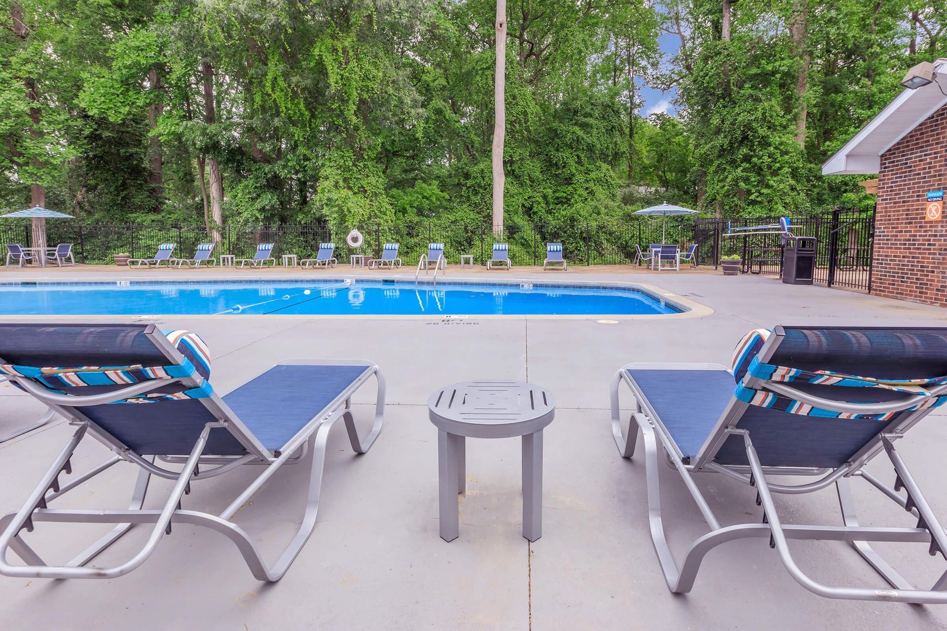 Two blue lounge chairs face a swimming pool. Trees and umbrellas in the background.