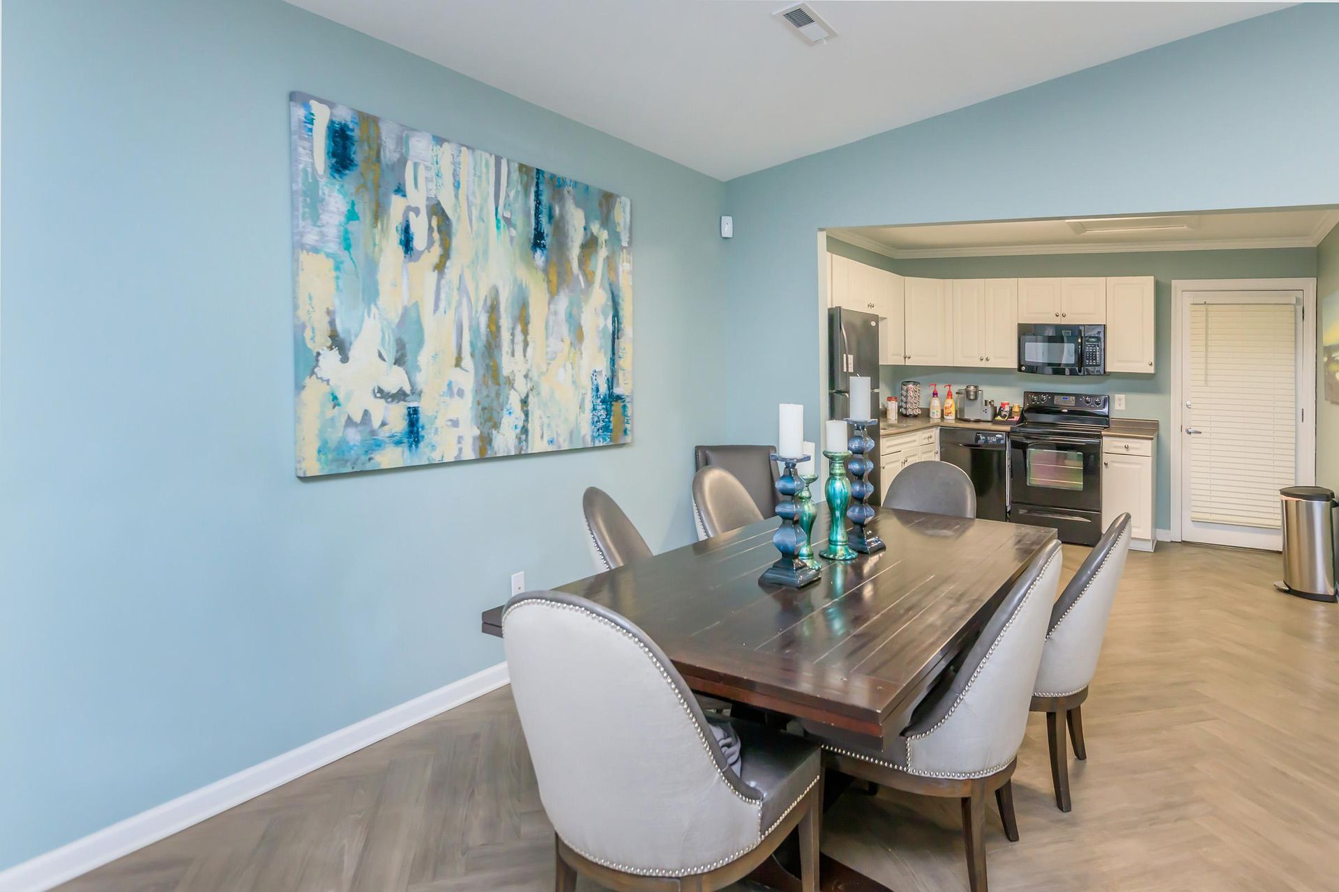 Dining area with a table, chairs, artwork, and a view into a kitchen. Light blue walls.