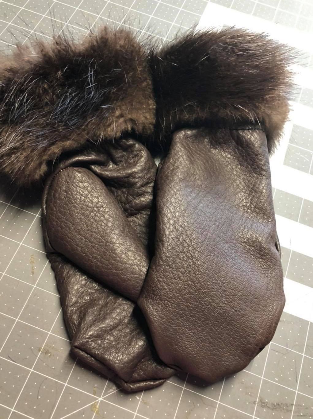 A pair of brown leather mittens with fur trim are sitting on a cutting board.