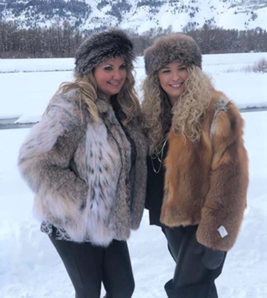 Two women wearing fur coats and hats are standing in the snow