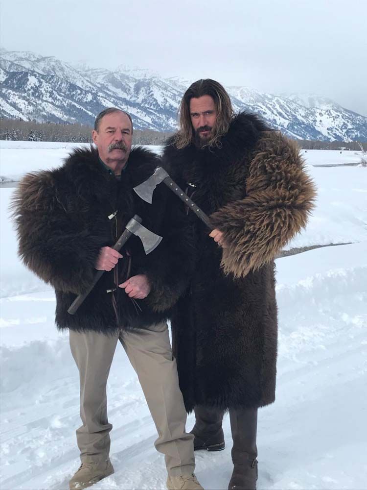 Two men in fur coats are standing in the snow holding axes