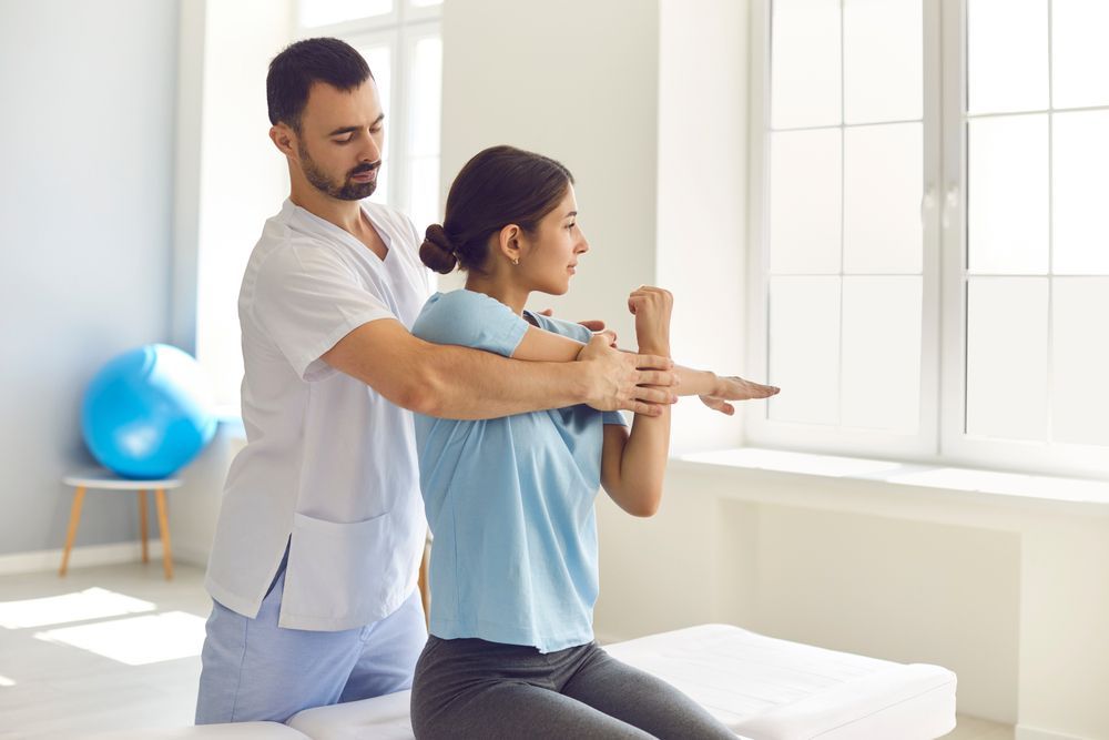 Physical therapist guides woman's arm stretch in a bright room with window.