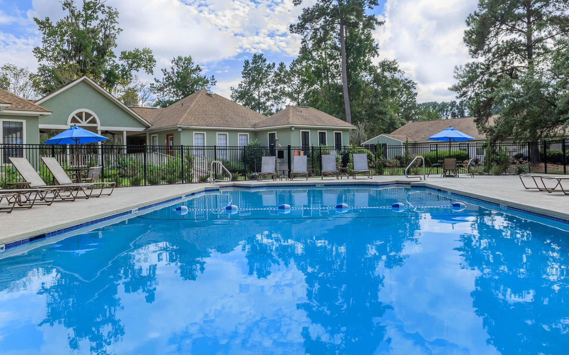 Swimming pool surrounded by lounge chairs, a fence, and apartments on a sunny day.