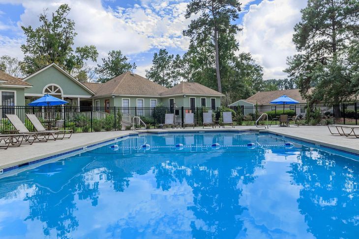 Swimming pool surrounded by lounge chairs, a fence, and apartments on a sunny day.