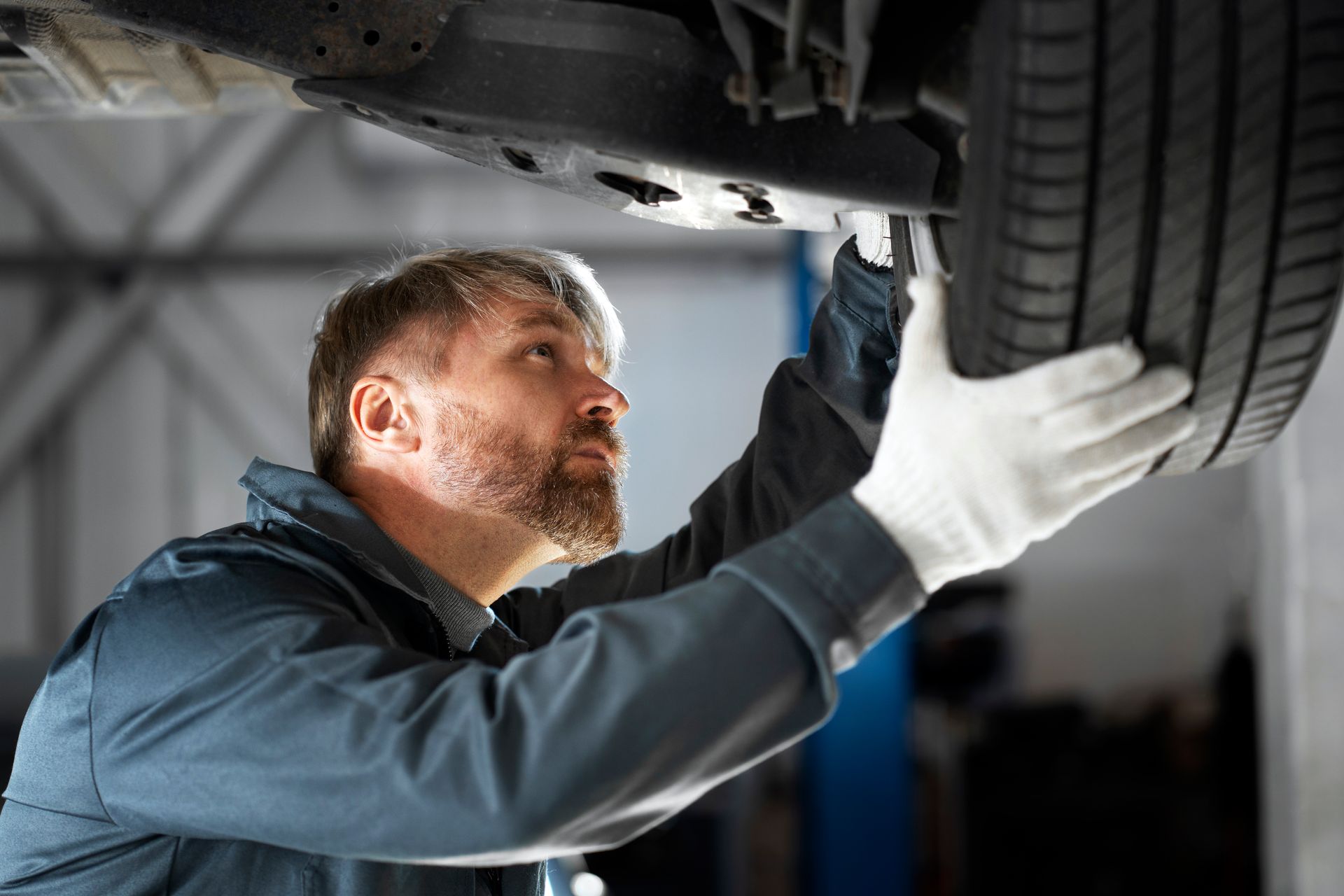 A mechanic in a uniform and gloves inspects the underside of a car lifted in a garage.