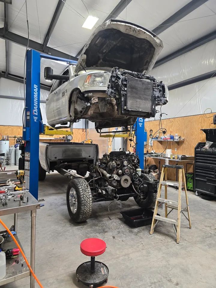 A white truck body is lifted on a two-post blue lift, separated from its chassis and engine in a professional garage.