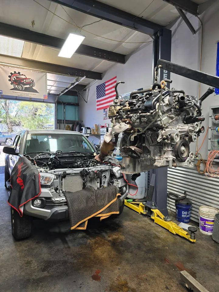 A vehicle’s engine hanging from a crane in an auto repair shop, with the open-hood truck parked nearby.
