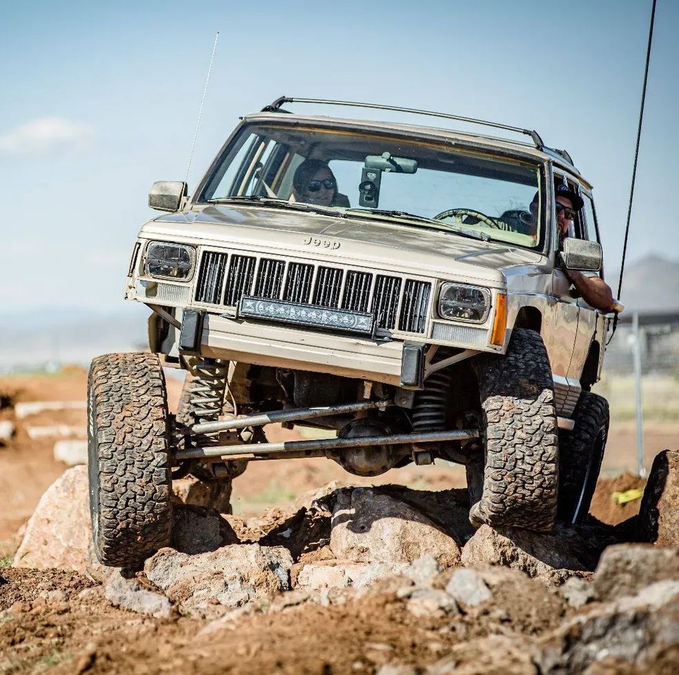 A beige Jeep Cherokee off-roading over rocky terrain, balanced with one front wheel lifted high in the air.