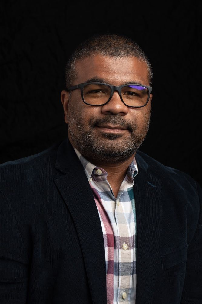 A man wearing glasses and a plaid shirt is standing in front of a black background.