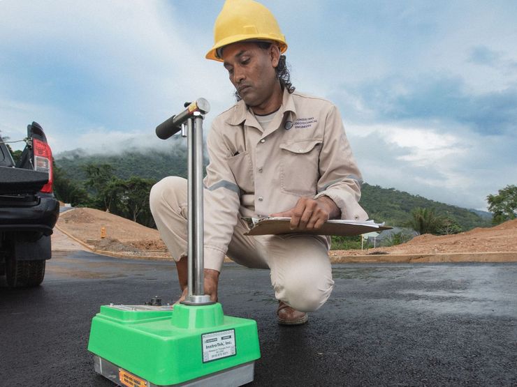 A man wearing a hard hat is kneeling down and looking at a clipboard.