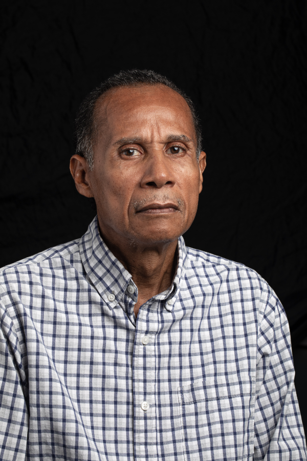 A man in a plaid shirt is standing in front of a black background.