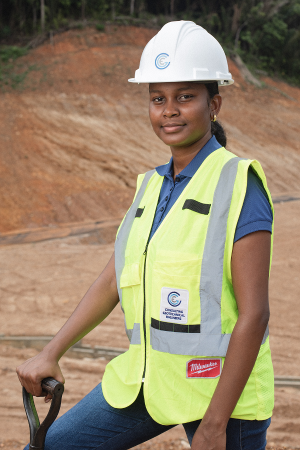 A woman wearing a hard hat and a yellow vest is holding a shovel.