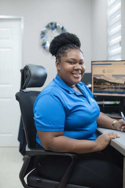 A woman in a blue shirt is sitting at a desk in front of a computer.