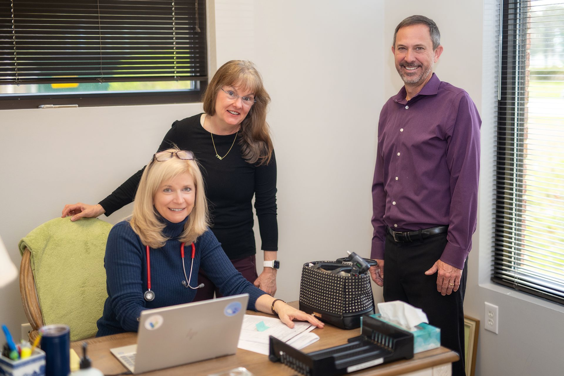 A woman is sitting at a desk with a laptop and two people are standing around her.