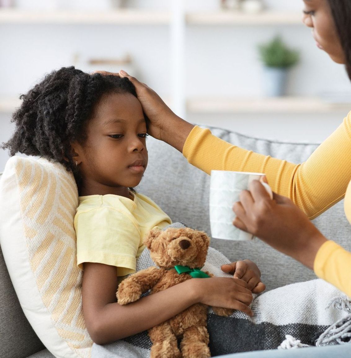 A little girl is laying on a couch with a teddy bear and a mother is holding her hand on the girl's forehead.