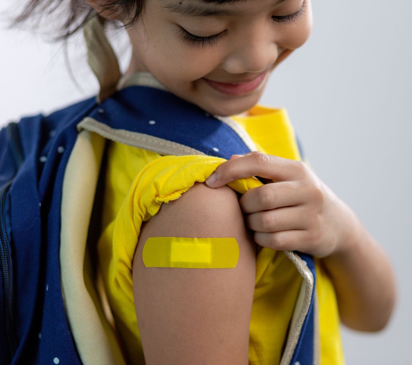 A young girl is wearing a backpack and yellow shirt. She is looking at her arm that has a yellow band-aid on it.