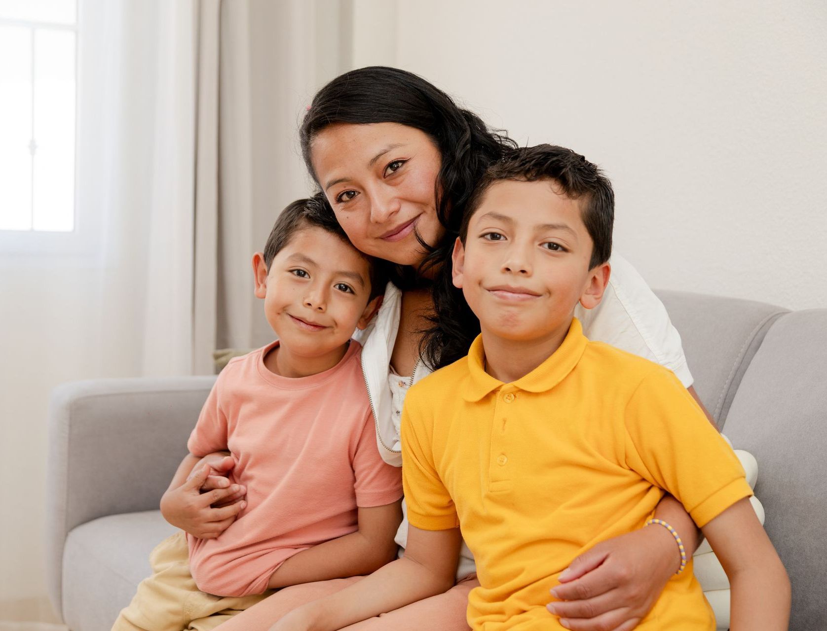 A mom has her arms around her two boys, sitting on a sofa. They are all smiling and looking at the camera.