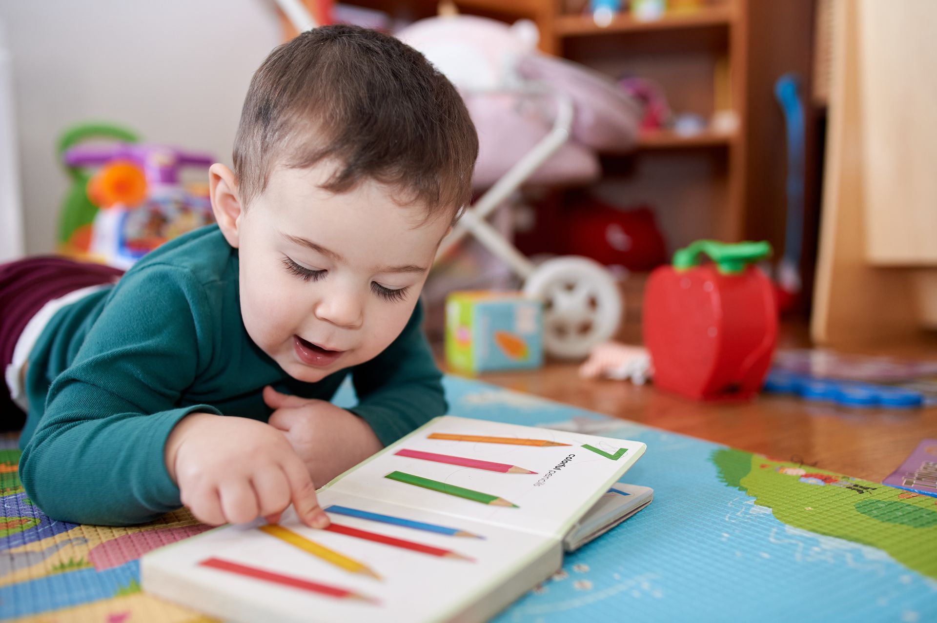 A young boy is lying on his stomach, reading a book. Toys are in the background.
