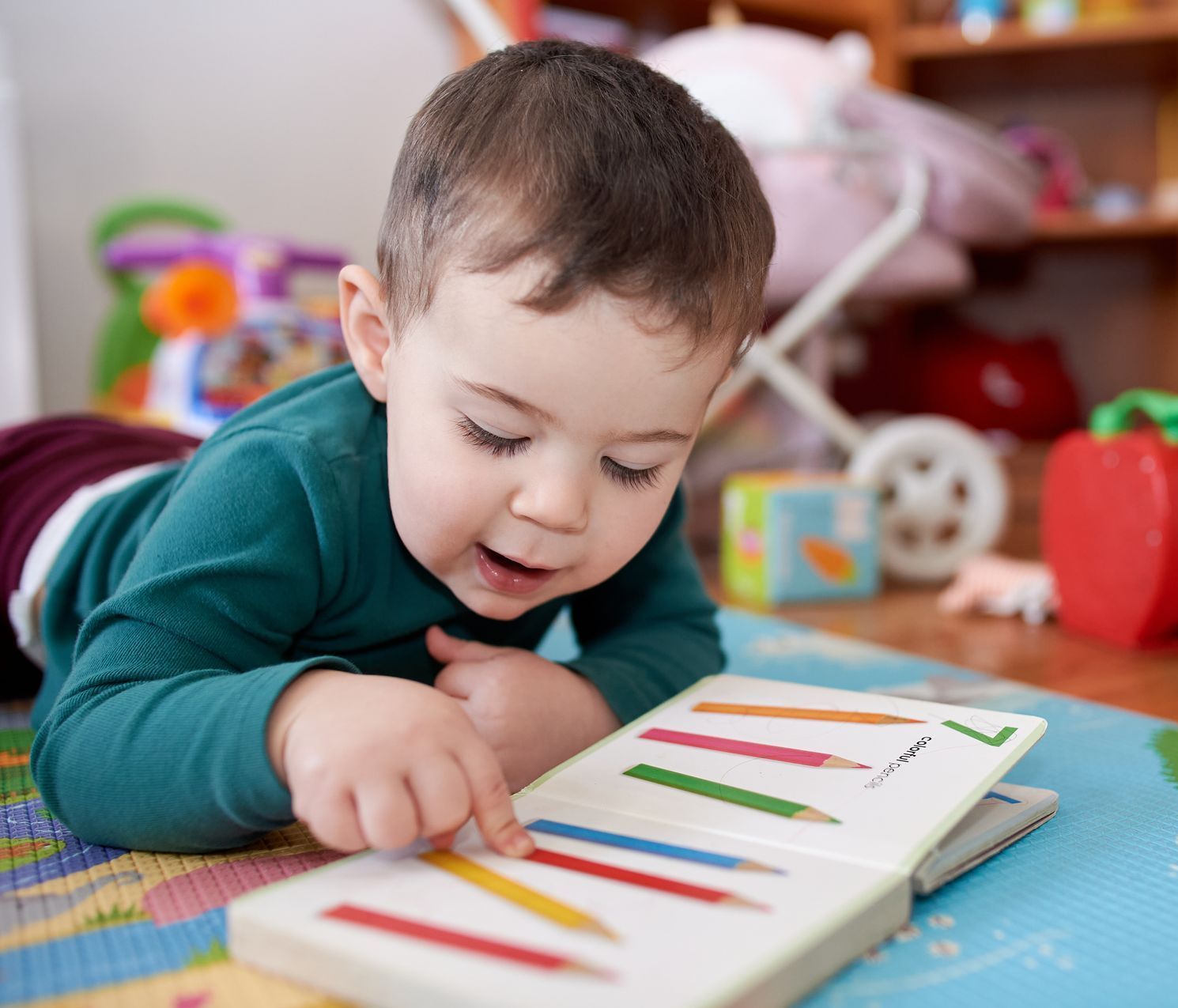 A little boy is laying on the floor reading a book with pencils on it.