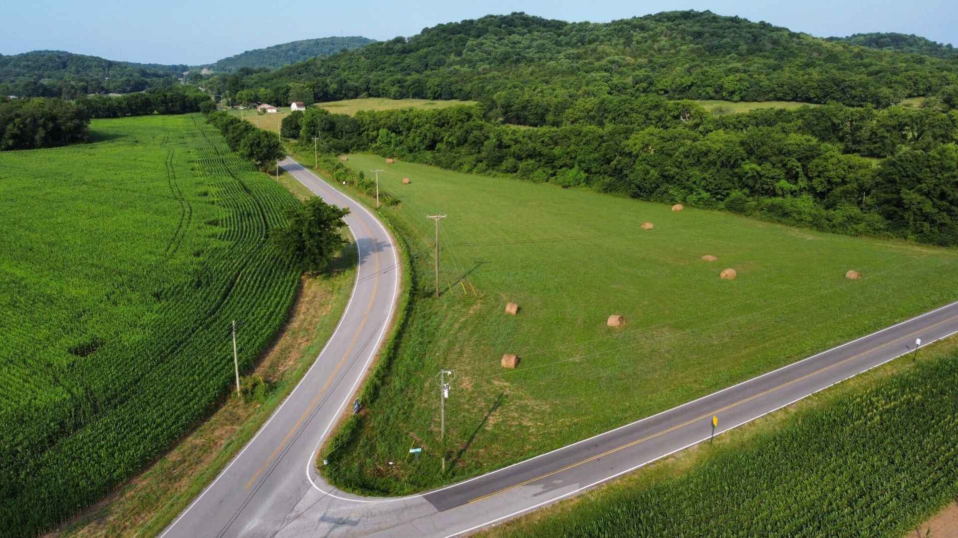 Winding road through green fields, bales of hay, and hills. Lush landscape with clear sky.