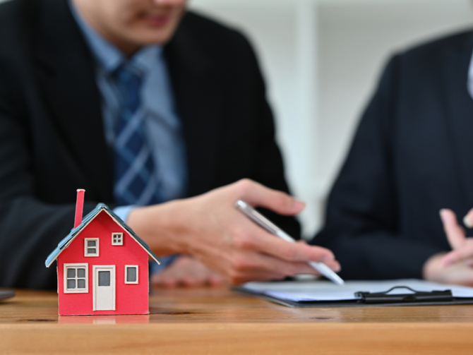 Red toy house on table, two men in suits discussing paperwork.