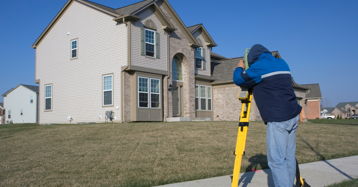 Surveyor using equipment in front of a two-story house, brown grass, blue sky.