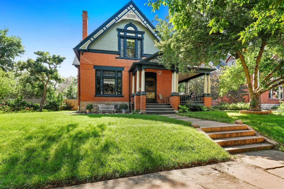 Victorian house with red brick exterior, porch, and green lawn under a blue sky.