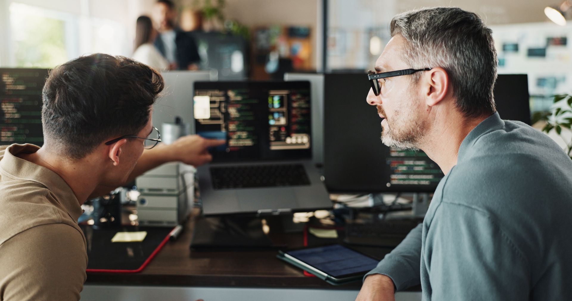 Two men at a desk, looking at a laptop screen, discussing a project. Office setting, focused expressions.